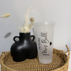 Puede incluir: Un vaso reutilizable de pl&aacute;stico transparente esmerilado con una pajita negra y una pegatina de vinilo negro que dice "FUTURE Mrs. Bull" con un peque&ntilde;o coraz&oacute;n negro. El vaso est&aacute; sobre una bandeja de mimbre trenzado con un jarr&oacute;n negro con flores secas blancas.