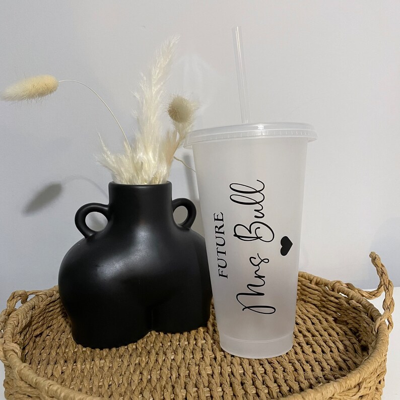 Puede incluir: Un vaso reutilizable de pl&aacute;stico transparente esmerilado con una pajita negra y una pegatina de vinilo negro que dice "FUTURE Mrs. Bull" con un peque&ntilde;o coraz&oacute;n negro. El vaso est&aacute; sobre una bandeja de mimbre trenzado con un jarr&oacute;n negro con flores secas blancas.