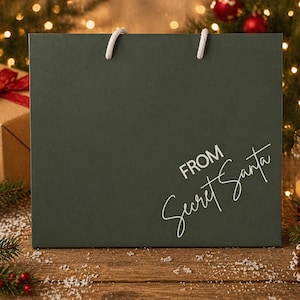 May include: A dark green gift bag with white rope handles, featuring the text "FROM Secret Santa" in a handwritten style. The bag is on a wooden surface, surrounded by Christmas decorations, including a lit candle, ornaments, and a gift box.