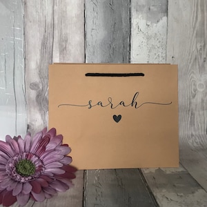 May include: A brown paper gift bag with a black handle and the name "sarah" in cursive script, with a black heart below. A purple flower is in the lower left corner. The bag is on a wooden background.