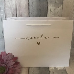 May include: White gift bag with a gold inscription that reads "nicola" with a small gold heart below the name.