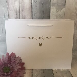 May include: White gift bag with a gold cursive name "emma" and a small gold heart.