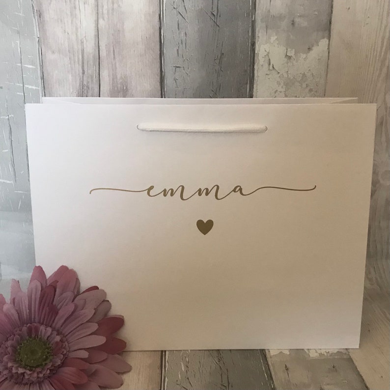 May include: White gift bag with a gold cursive name "emma" and a small gold heart.
