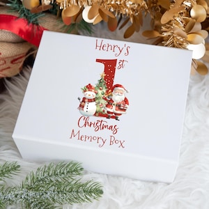 May include: White box with a red and white design featuring a snowman, Santa Claus, and a Christmas tree. The box says "Henry's 1st Christmas Memory Box".