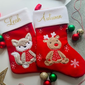 Peut inclure: Deux chaussettes de Noël rouges avec des revers blancs. La chaussette de gauche a un renard avec une écharpe rouge brodée dessus et le nom "Leah" sur le revers. La chaussette de droite a un renne avec une écharpe rouge brodée dessus et le nom "Autumn" sur le revers.