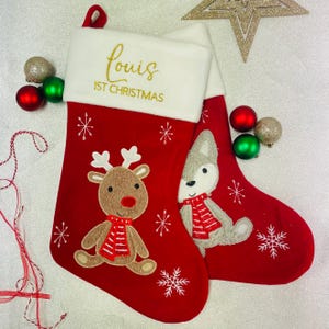 May include: Two red Christmas stockings with white cuffs. One stocking features a reindeer with a red and white scarf, the other a wolf with a similar scarf. The text "Louis 1st Christmas" is embroidered in gold on the cuff.