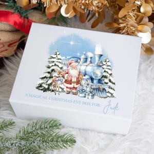 May include: A white gift box featuring a blue and white illustration of Santa Claus, a reindeer, and a train in a snowy woodland setting. The text "A magical Christmas Eve box for: Jude" is printed on the box.