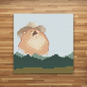 May include: Cross-stitch picture of a light brown animal wearing a cowboy hat. The image shows the animal against a light blue sky and a green and brown landscape. The artwork is framed and displayed on a wooden surface.