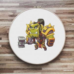 May include: Cross stitch pattern of a green orc wearing gold and purple armor and holding a hammer. The orc has a serious expression on its face.