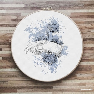 May include: A cross-stitch embroidery design featuring a blue and white animal skull with blue flowers. The skull is surrounded by a blue and white pixelated background.