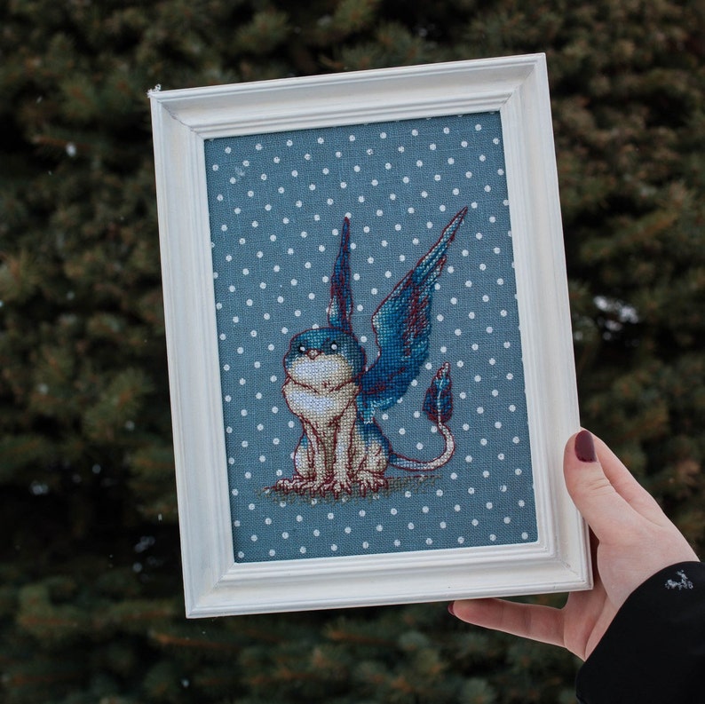 May include: A framed cross-stitch embroidery of a blue and white winged creature with a long tail, sitting on a blue and white polka dot background.