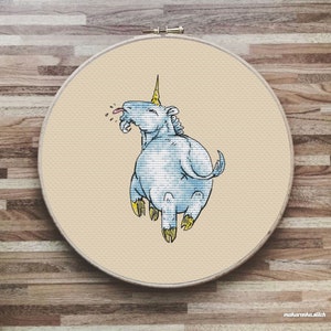 Puede incluir: Un unicornio azul con un cuerno amarillo y una lengua rosa que sobresale está bordado en una tela beige. El unicornio está rodeado de un círculo blanco.