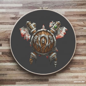 Könnte beinhalten: Ein Kreuzstichmuster eines Schildes mit zwei Äxten, mit einem braunen Holzschild mit silbernem Rand und einem grünen Edelstein an der Spitze jeder Axt. Der Schild hat ein stilisiertes 'H'-Symbol in der Mitte.