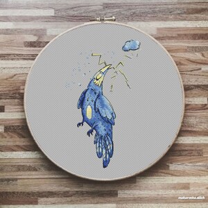 Puede incluir: Un pájaro azul con detalles amarillos está bordado en un lienzo redondo gris. El pájaro canta en un rayo amarillo. Una pequeña nube azul está sobre el pájaro.
