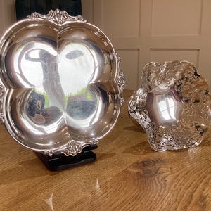 May include: Two silver serving dishes, one with a scalloped edge and the other with a more ornate design. Both dishes are polished and reflect light.