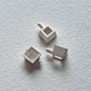 Könnte beinhalten: Drei silberfarbene Metall-Anhänger in Form eines Quadrats mit einer kleinen Öffnung in der Mitte. Die Anhänger haben eine kleine Schlaufe an einer Seite zum Anbringen an einer Halskette oder einem Armband.