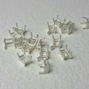 Puede incluir: Un grupo de engastes de metal plateado para piedras preciosas. Los engastes tienen forma de rectángulo con esquinas redondeadas y cuatro garras a cada lado.