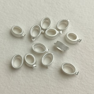 May include: A collection of small, oval-shaped silver-coloured jewellery findings. Each finding has a loop on one side, and a small rectangular tab on the other. The findings are scattered on a light-coloured surface.
