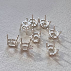 May include: A group of ten silver-toned metal oval-shaped settings for jewellery making. The settings have four prongs each and are designed to hold a gemstone or other embellishment.