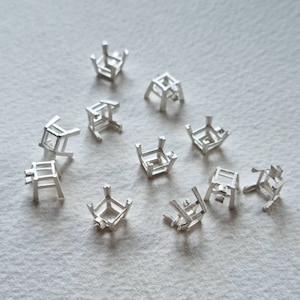 Puede incluir: Un conjunto de 10 engastes de metal plateado para montar piedras preciosas. Los engastes están en varias posiciones y ángulos, mostrando su diseño intrincado.