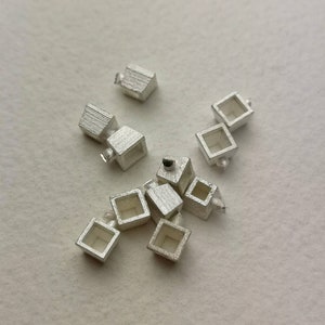 May include: A group of nine silver-coloured, hollow, square-shaped charms with a small loop at the top. The charms are scattered on a white surface.