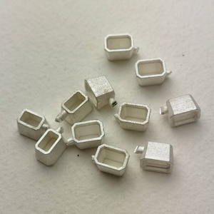 Può includere: Un gruppo di dieci ritrovamenti metallici color argento, ognuno con forma rettangolare e un piccolo foro nella parte superiore. I ritrovamenti sono sparsi su una superficie bianca.