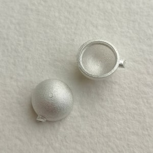 Puede incluir: Dos cuentas de metal plateado, una es una esfera sólida y la otra es una esfera hueca con un pequeño agujero en el lateral.