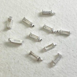 Puede incluir: Una colección de pequeños componentes metálicos plateados, posiblemente hallazgos de joyería o conectores, dispersos sobre una superficie clara. Cada pieza tiene un cuerpo rectangular con una pequeña sección que sobresale en un extremo.