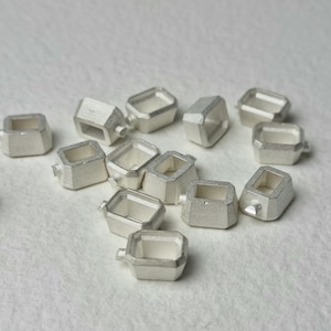May include: A group of 12 silver-toned metal charms, each shaped like a small, open box with a square opening. The charms are scattered on a white surface.