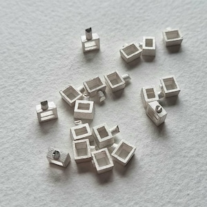May include: A scattering of small, silver-coloured metal components. Each piece has a rectangular box shape with a small, cylindrical post extending from one side. The components are scattered on a light-coloured surface.