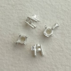 May include: Four silver metal settings for mounting gemstones. The settings are in various shapes, including a round, a square, and two prongs.