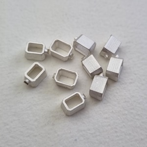 Puede incluir: Un grupo de diez biseles rectangulares de metal de color plateado con un pequeño agujero en la parte superior. Los biseles están dispersos sobre una superficie blanca.