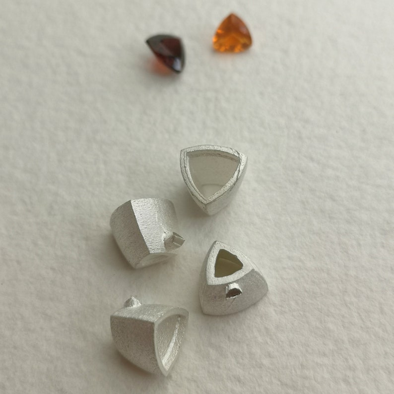 May include: Four silver triangular settings for gemstones, with two orange and one brown gemstones on a white surface.