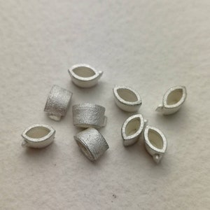 Könnte beinhalten: Eine Sammlung silberfarbener Metallperlen, einige in Blattform und andere zylindrisch. Die Perlen sind klein und haben eine glatte, polierte Oberfläche.