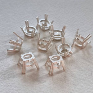 May include: A group of ten silver-colored metal settings for oval-shaped gemstones. The settings have six prongs each and are designed to hold a stone in place.