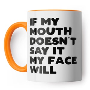 May include: White ceramic coffee mug with an orange rim and handle. The mug has black text that reads "If my mouth doesn't say it my face will."