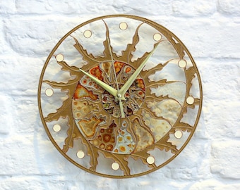 Puede incluir: Un reloj de pared redondo de color dorado con un diseño decorativo en forma de sol. La esfera del reloj presenta un patrón circular multicolor sobre fondo blanco. Las manecillas del reloj son blancas.