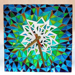 May include: A square clock with a white snowflake design in the center. The clock face is surrounded by a mosaic of blue, green, and brown triangles.