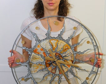 Puede incluir: Un reloj de pared grande y redondo con un diseño único. El reloj presenta un marco de metal plateado con un centro detallado en forma de fósil en tonos marrones, naranjas y crema. Acentos dorados marcan las horas. Las manecillas del reloj son de madera.