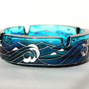 Puede incluir: Un cenicero de vidrio azul con un diseño de olas y montañas en vitral. El cenicero tiene un borde plateado y un diseño blanco.