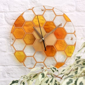 Puede incluir: Un reloj redondo con un diseño de panal en tonos amarillos y naranjas. El reloj tiene una aguja de madera y agujas negras. Pequeñas abejas están dispersas alrededor del diseño de panal.