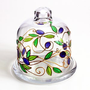 May include: A glass cloche with a gold, green, and blue painted design of leaves and berries. The cloche has a clear glass base.