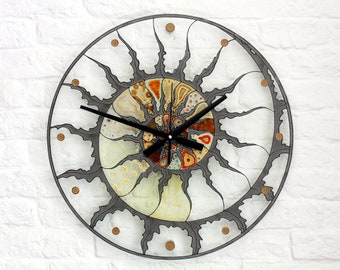 Puede incluir: Un reloj de pared grande, redondo, de metal gris con un diseño de rayos de sol. La esfera del reloj está hecha de vidrio y presenta un patrón abstracto colorido en tonos marrones, naranjas y amarillos. Las manecillas del reloj son negras.