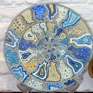 Puede incluir: Un plato redondo de cerámica con un diseño abstracto azul, blanco y dorado. El diseño presenta patrones en espiral y puntos.