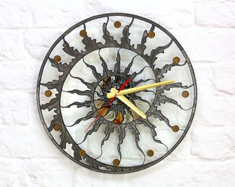 Puede incluir: Un reloj circular con un diseño de rayos de sol. La esfera del reloj está hecha de vidrio transparente con un marco de metal plateado. Las manecillas del reloj son rojas y doradas. El reloj está puesto a las 12:00.