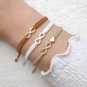 Könnte beinhalten: Drei Freundschaftsarmbänder mit Unendlichkeitszeichen und einem Herz-Anhänger. Die Armbänder sind aus braunem, weißem und beigefarbenem Kordel gefertigt.