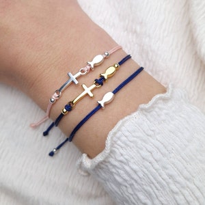 Könnte beinhalten: Drei Armbänder mit Anhängern an einem Handgelenk. Ein Armband ist pink mit einem silbernen Kreuzanhänger, eines ist dunkelblau mit einem goldenen Kreuzanhänger und eines ist dunkelblau mit einem goldenen Fischanhänger.