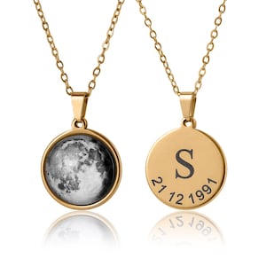 Puede incluir: Collar con colgante dorado con un dije redondo. Un lado muestra una imagen detallada en escala de grises de la luna. El otro lado tiene la letra "S" y la fecha "21.12.1991" grabada. El collar cuelga de una delicada cadena dorada.