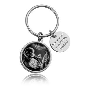 May include: Silver keychain with a round pendant featuring a black and white sonogram image. A smaller, circular charm is engraved with the words "Now you can carry me too, Daddy!"
