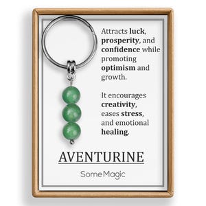 May include: Aventurine gemstone keychain with three green beads. The keychain is attached to a card with text that reads "Attracts luck, prosperity, and confidence..." and "AVENTURINE SomeMagic". The frame is a light brown color.